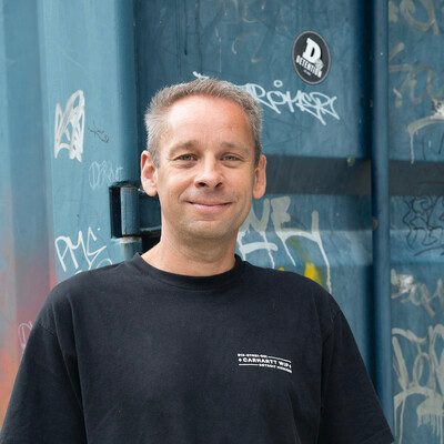 Joost Faassen in a black t-shirt, smiling, in front of a blue steel door covered in graffiti.
